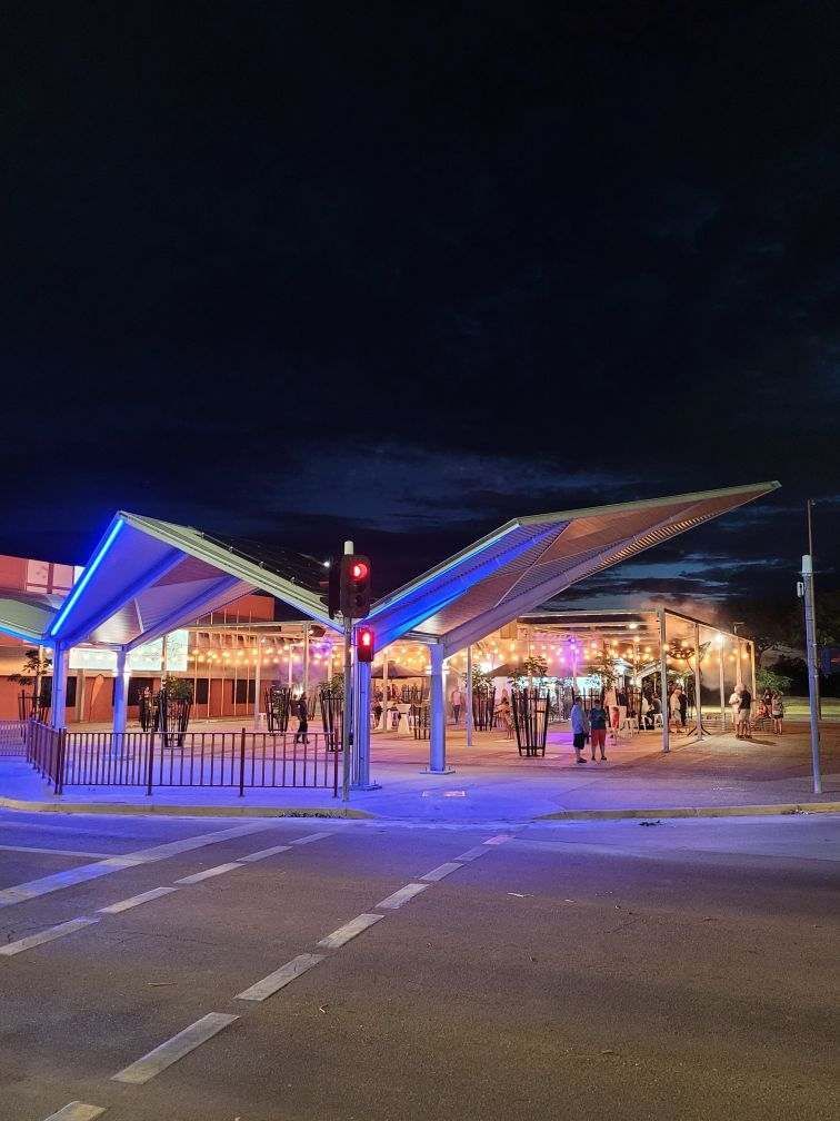 Illuminated modern canopy over a gated plaza at night, with colorful blue and pink lights and a few people walking