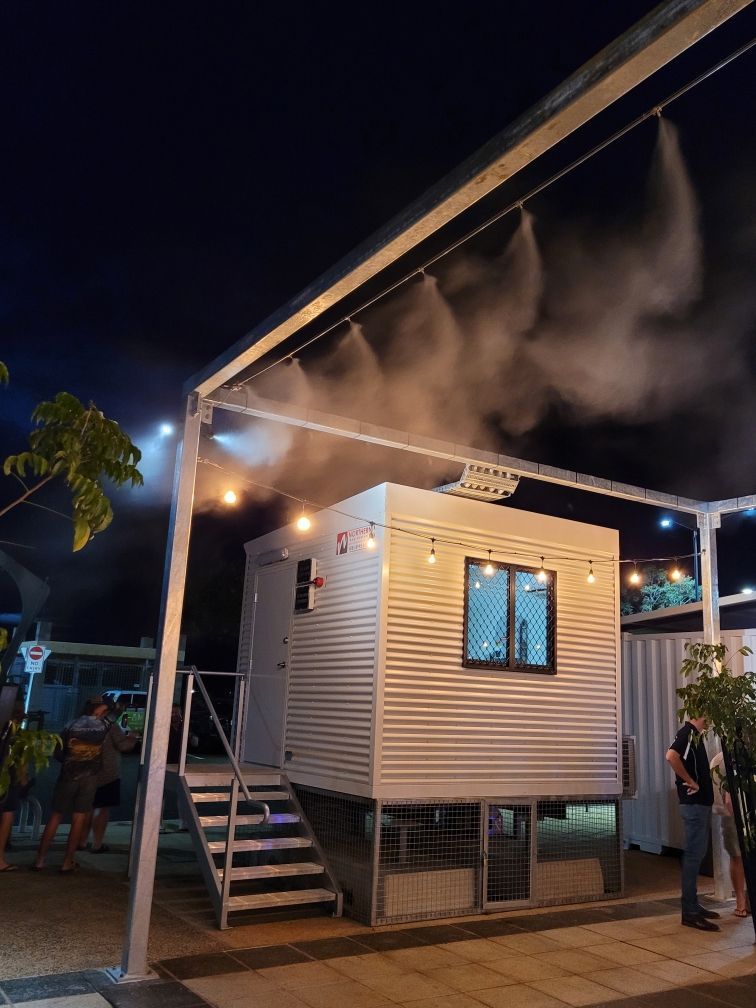 Small elevated house at night with mist from roof sprayers and people standing nearby