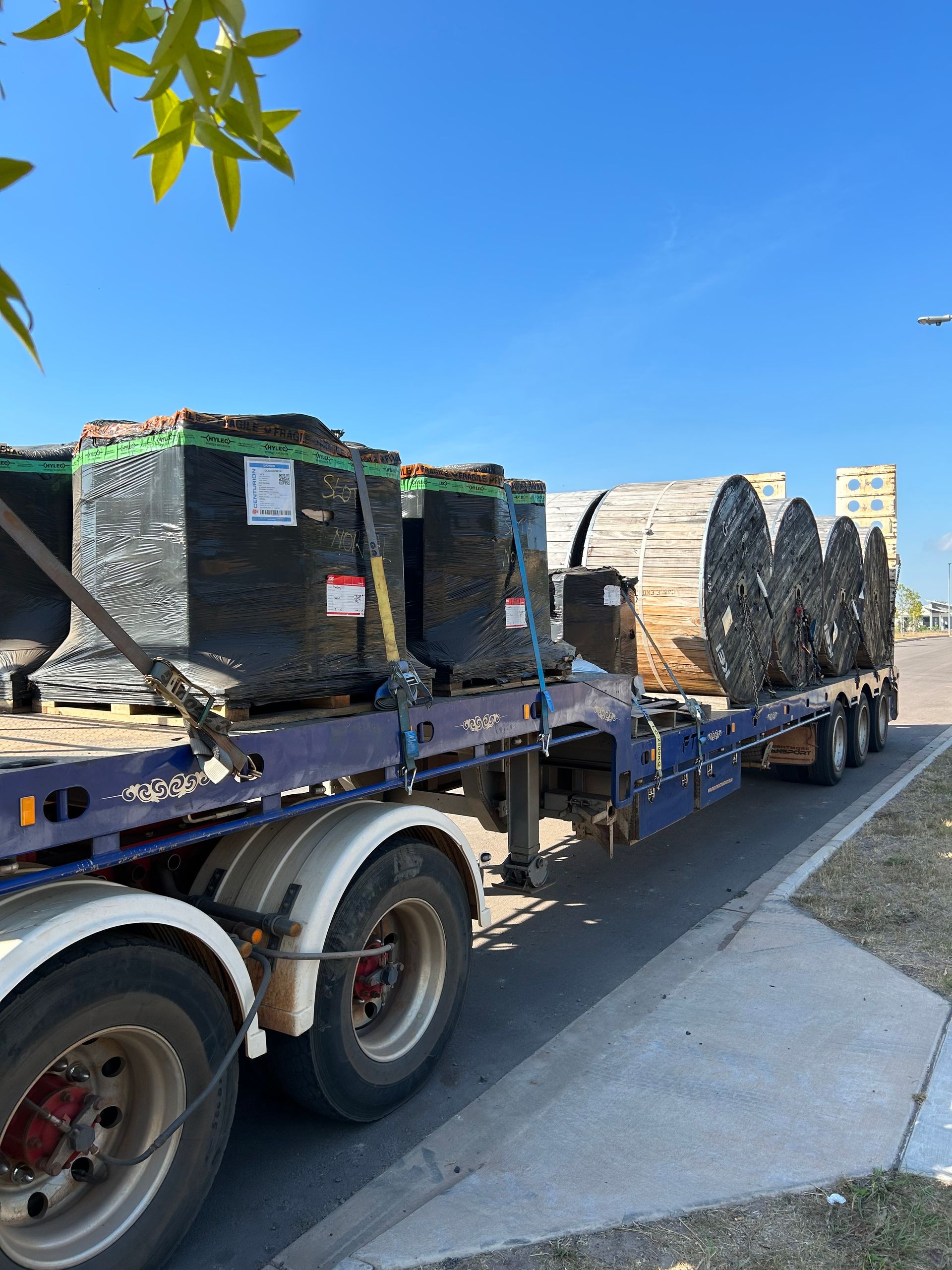Flatbed trailer carrying stacked black crates and large spools parked along a sunny roadside