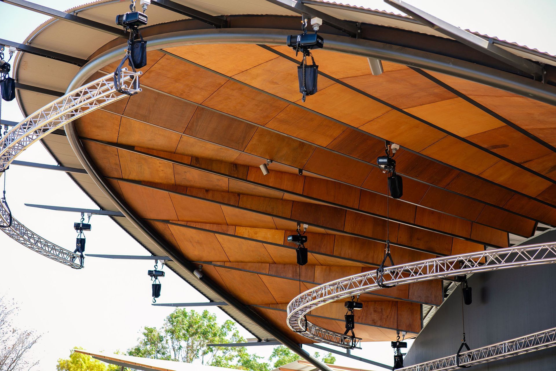 Curved wooden canopy with black metal truss lighting fixtures overhead outdoors