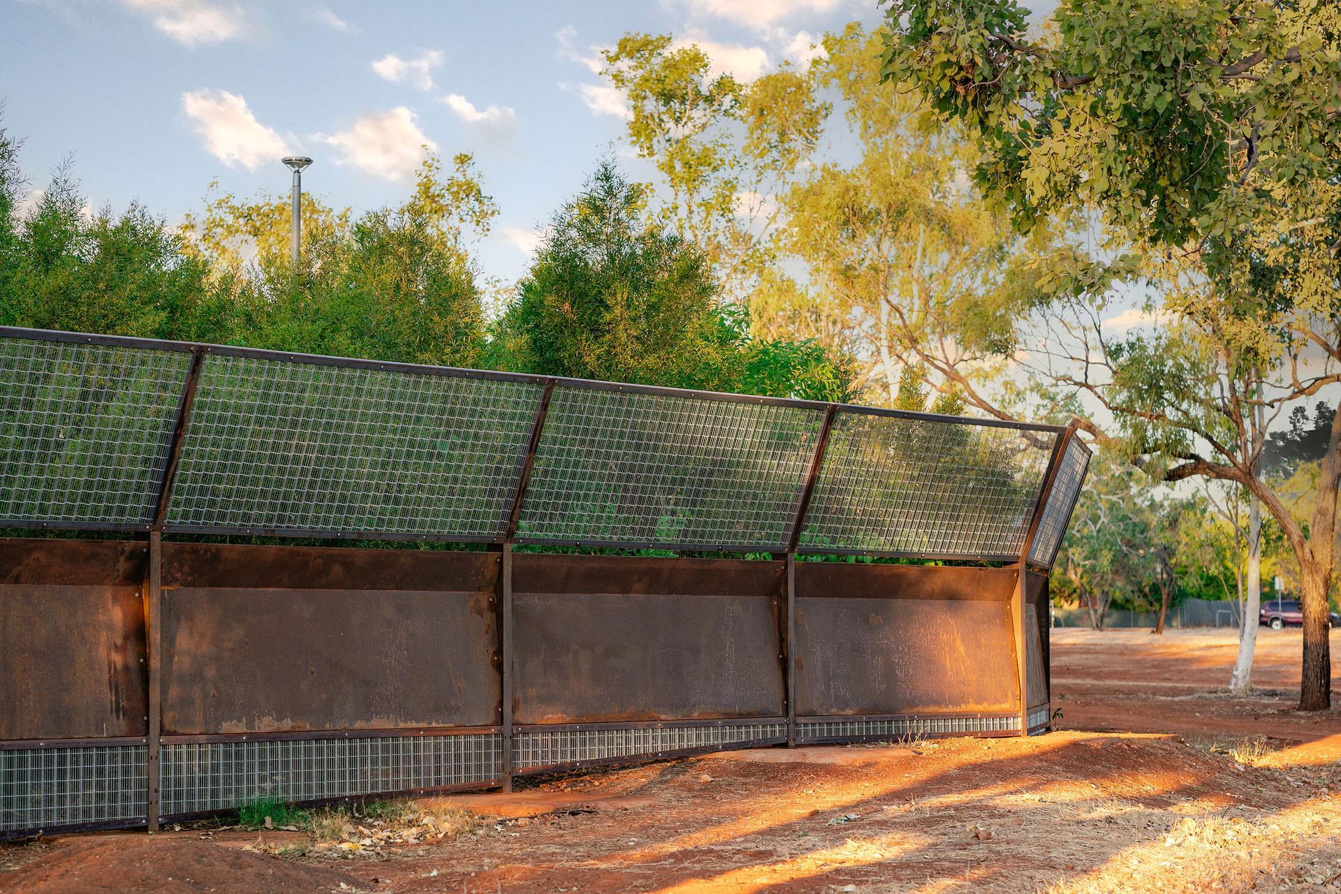 Long shelter with green roof panels in a sunlit park, surrounded by trees and dry ground