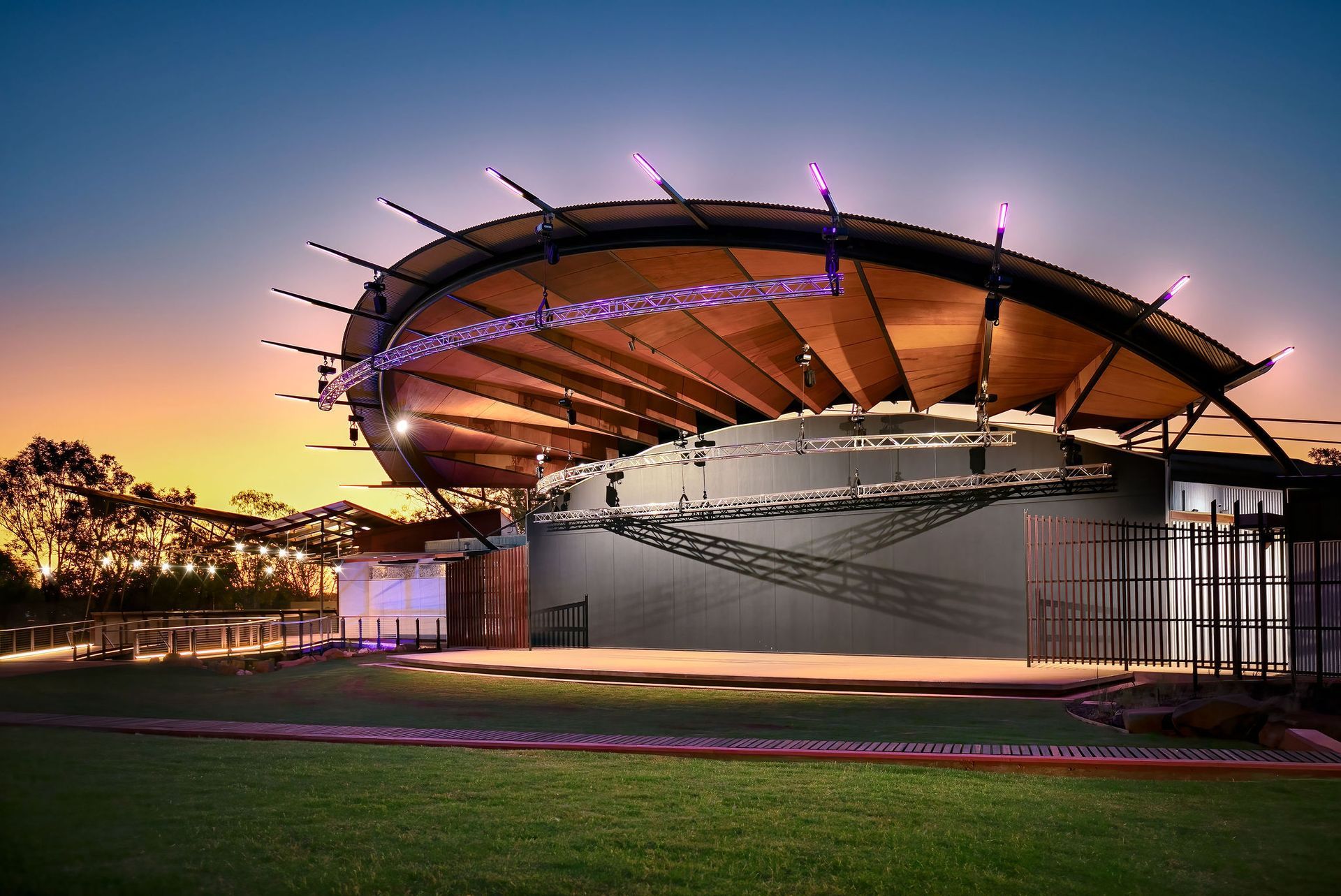 Modern outdoor pavilion with curved roof at sunset, lit with purple accents over a grassy field
