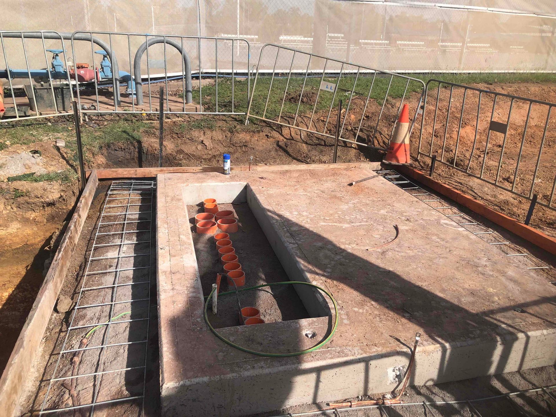 Construction site with a concrete pit, exposed rebar, and orange safety cones in a fenced area