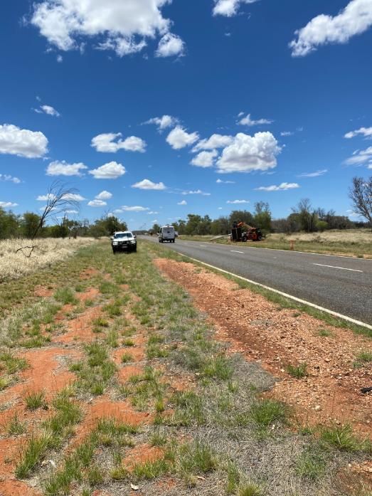 Rural roadside with parked vehicles, grassy median, and blue sky with scattered clouds.