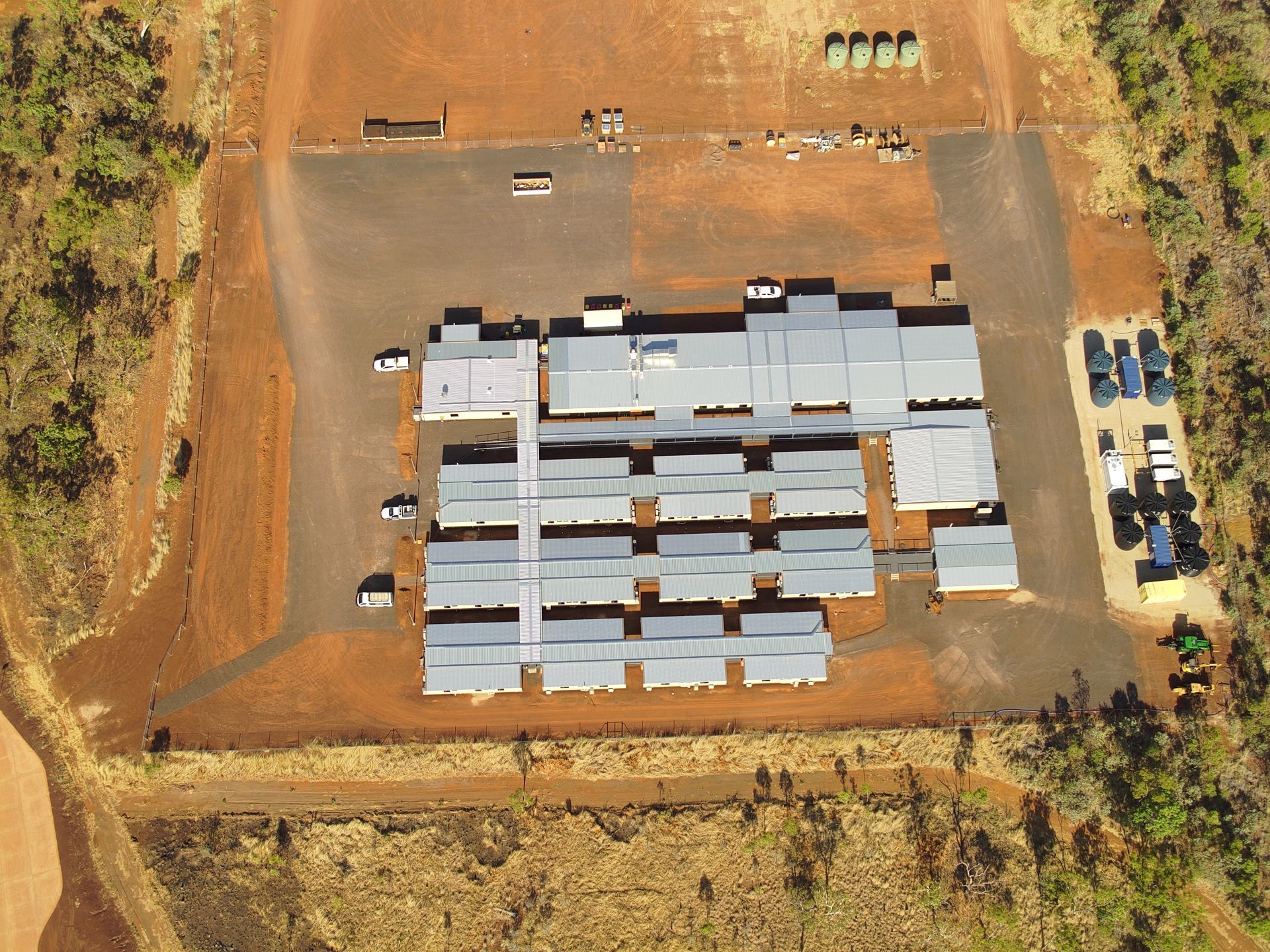 Aerial view of a construction site with rows of gray-roofed buildings on red dirt, surrounded by vehicles.