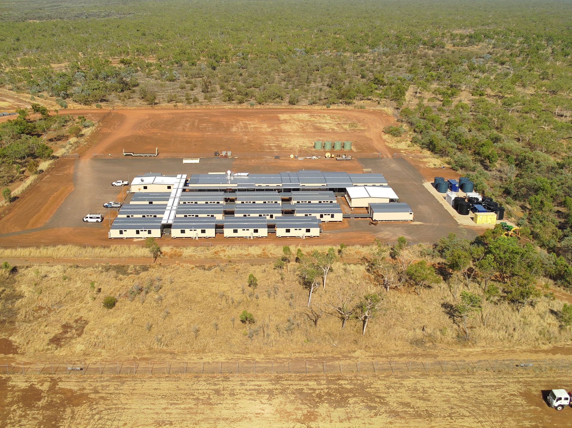 Aerial view of a remote building complex in a dry, wooded landscape, with dirt roads and parked vehicles.
