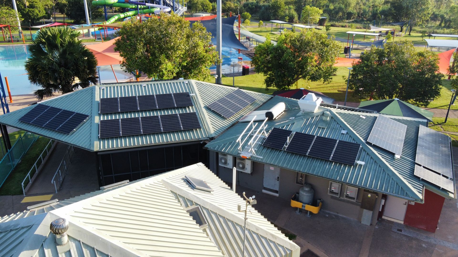 Aerial view of a modern building with green metal roofs and solar panels, surrounded by trees and a parking lot