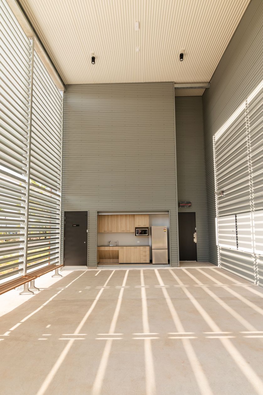 Empty modern room with tall louvered windows and a small kitchen area at the back, lit by bright sunlight.