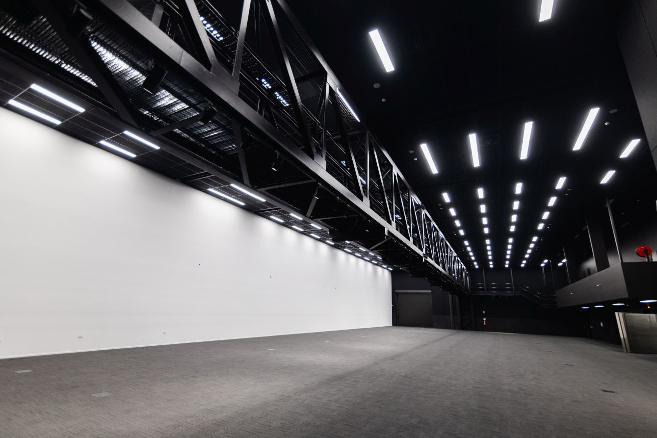 Empty indoor exhibition hall with a gray floor, white wall, and rows of bright ceiling lights