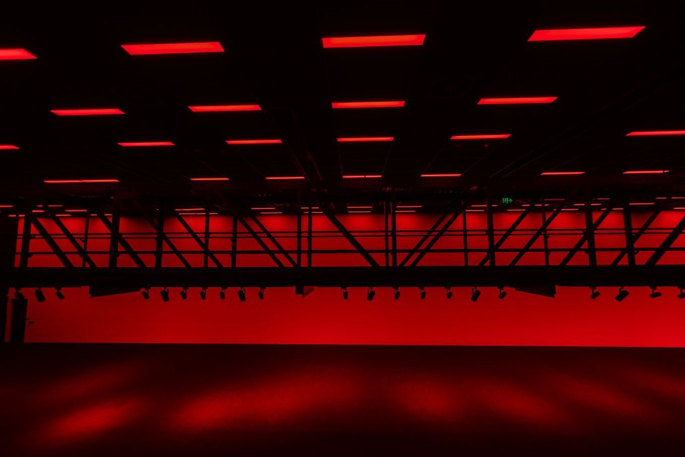 Abstract red-lit stage with overhead lights and silhouetted truss structure, fading into darkness.