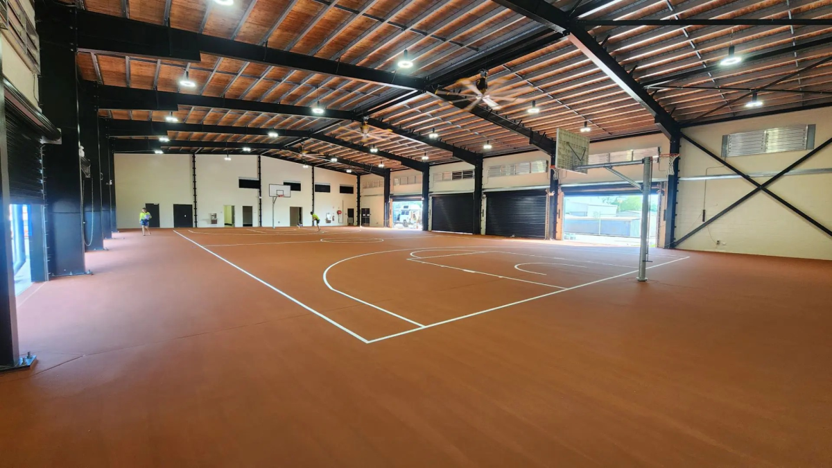 Empty indoor sports hall with orange flooring and white court markings under a high metal roof