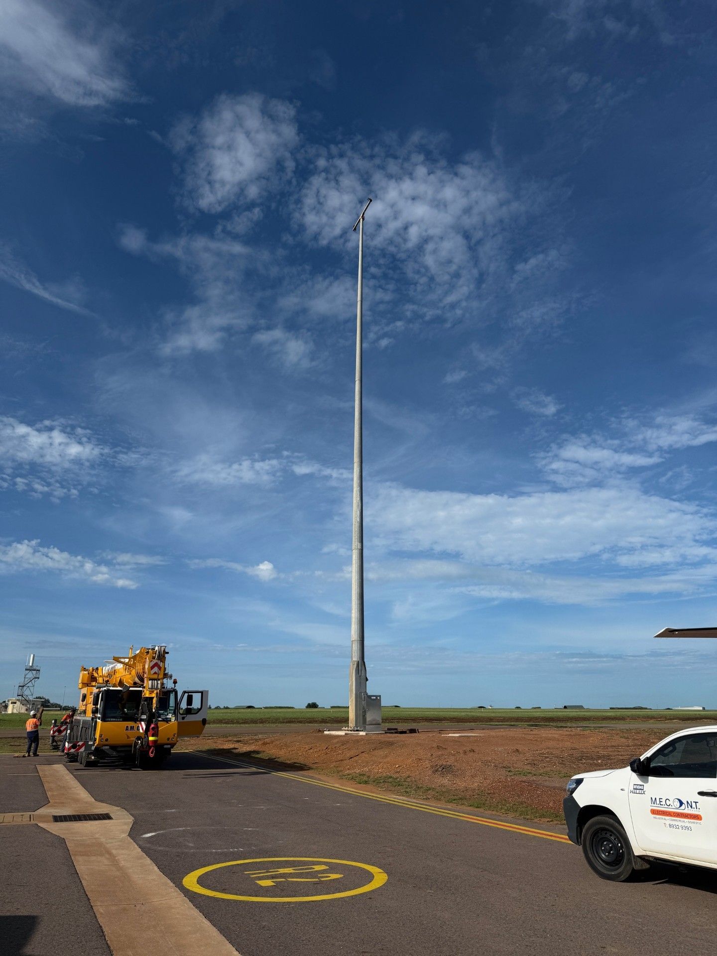 Tall light pole under a blue cloudy sky, with vehicles parked on a paved roadside.