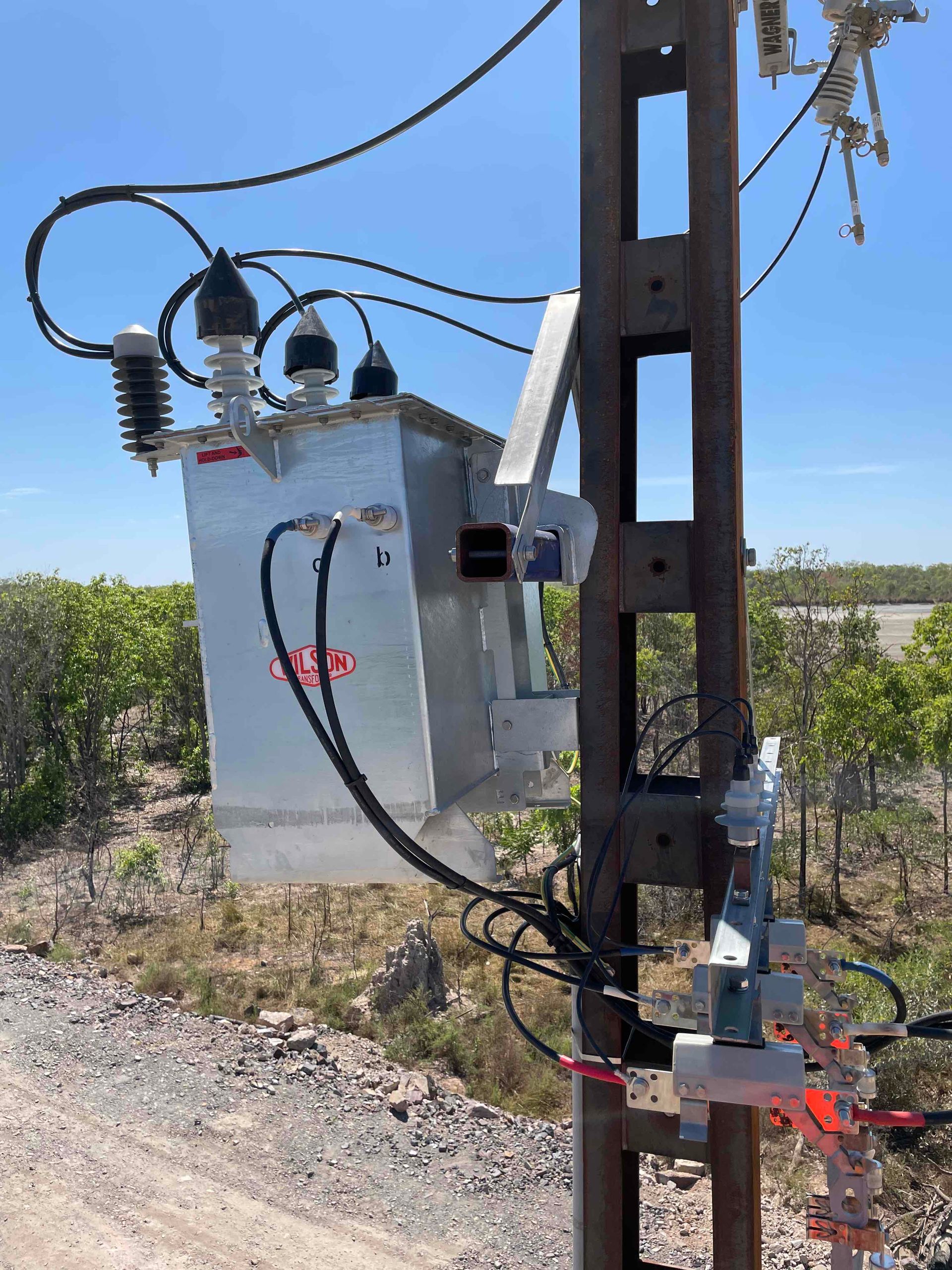 Electrical transformer mounted on a utility pole in a dry roadside field under a blue sky