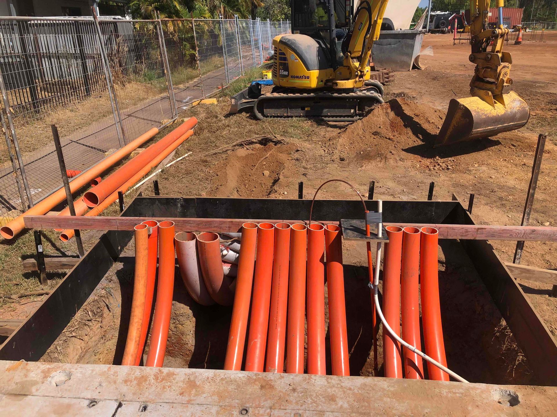Utility trench with orange conduit pipes and excavators at a roadside construction site.