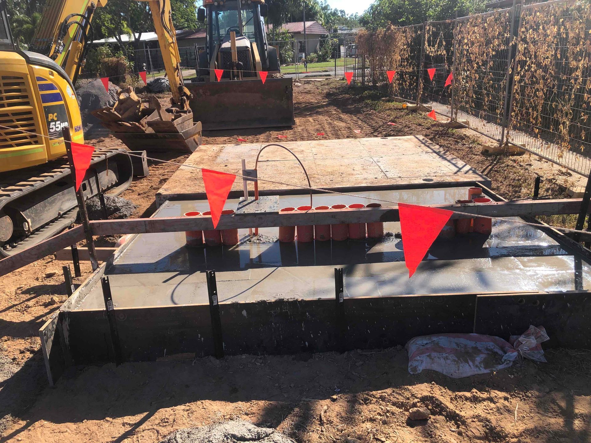 Construction site with a small excavator, concrete slabs, and red safety flags along a trench