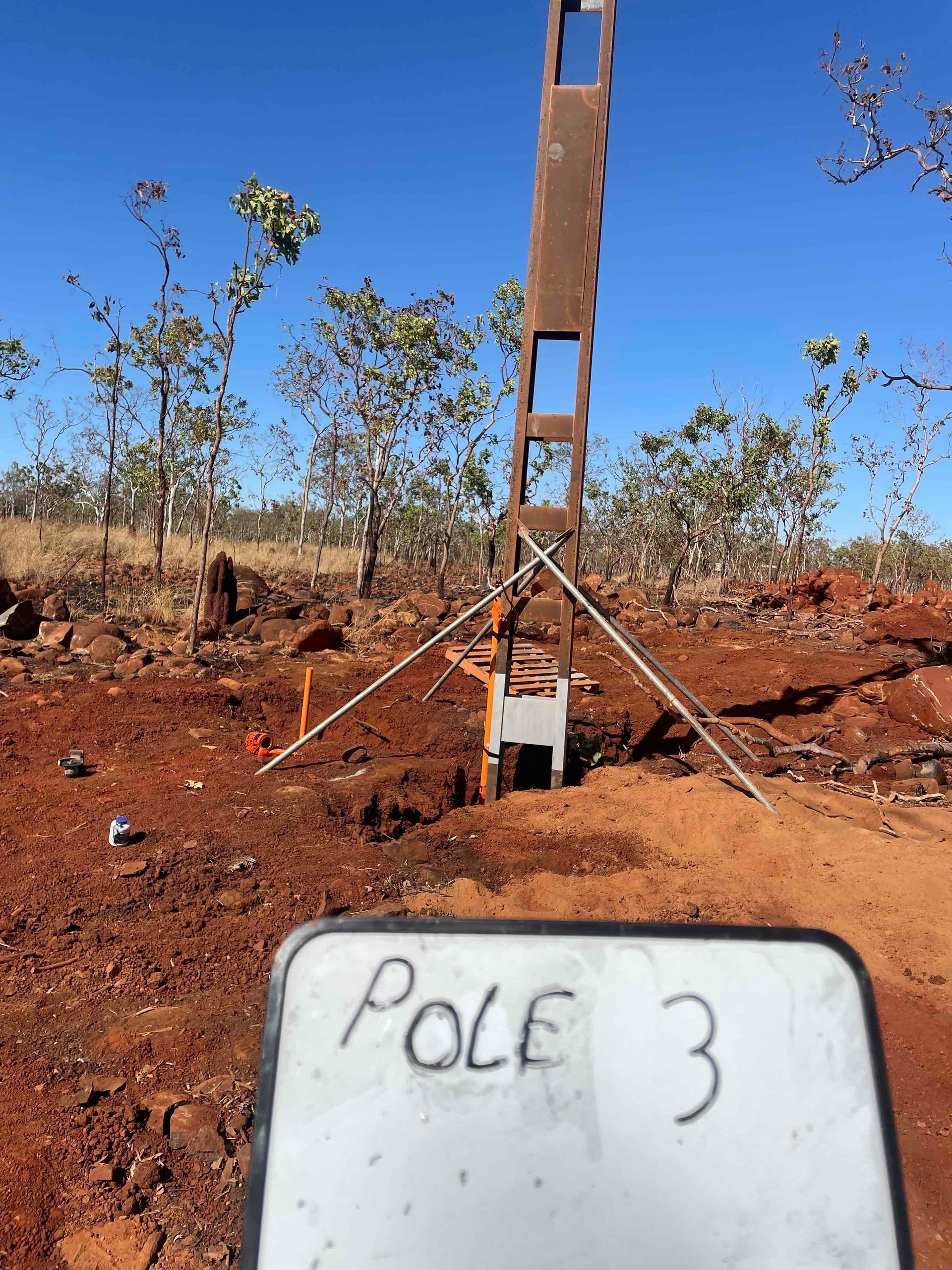 Pole 3 marker on a reddish dirt field with a tall metal pole and sparse trees in the background