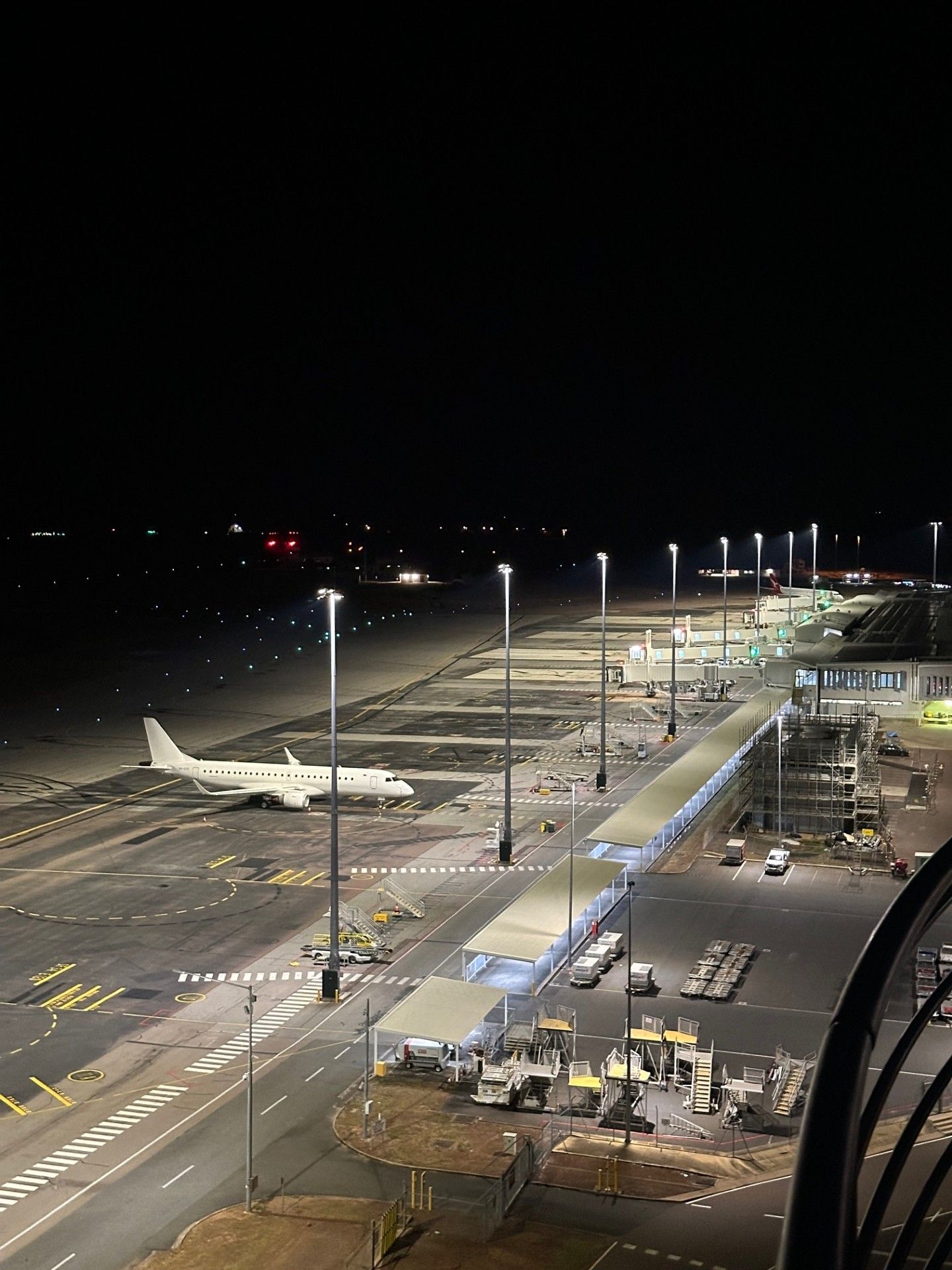 Airplane parked at a brightly lit airport gate at night, with terminal buildings and service vehicles nearby
