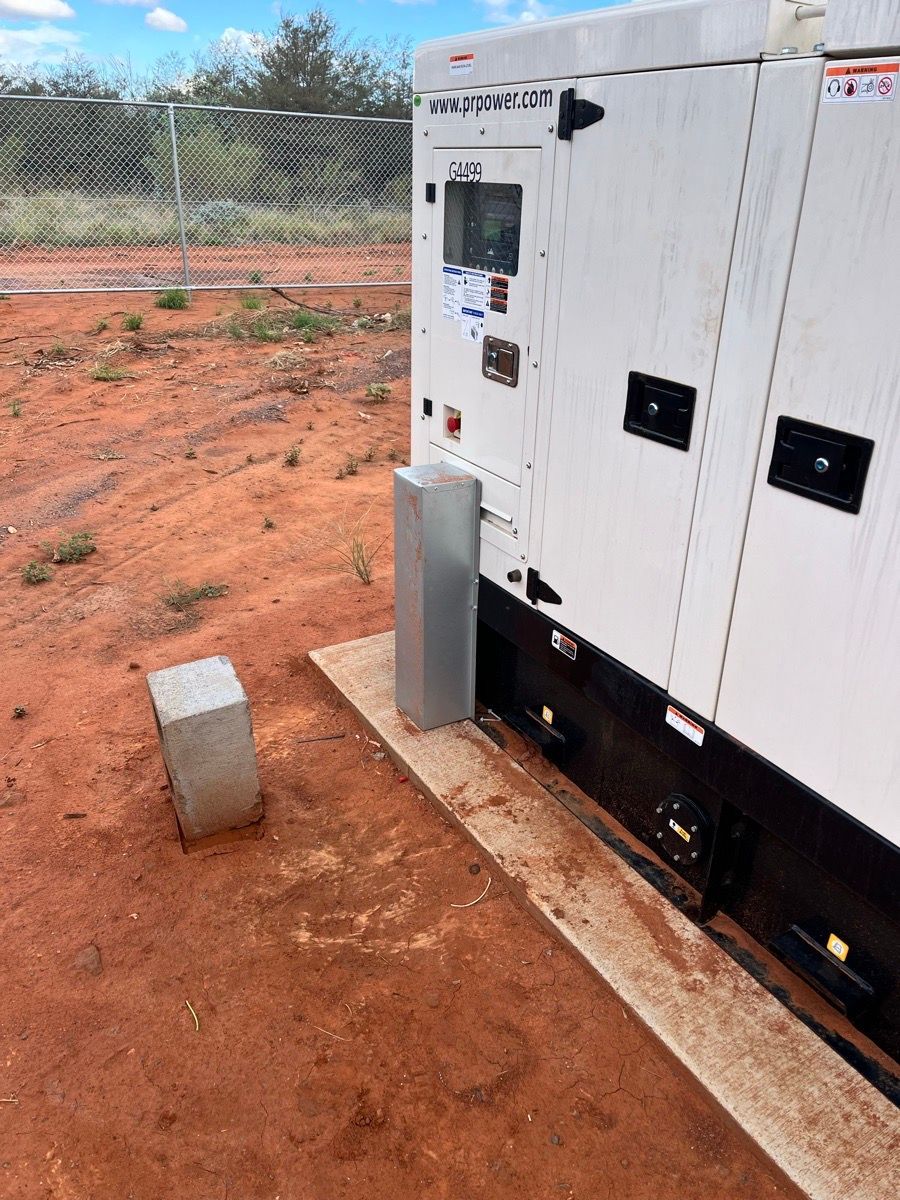 White utility trailer beside a chain-link fence on red dirt, with concrete blocks and a gray cylinder nearby.