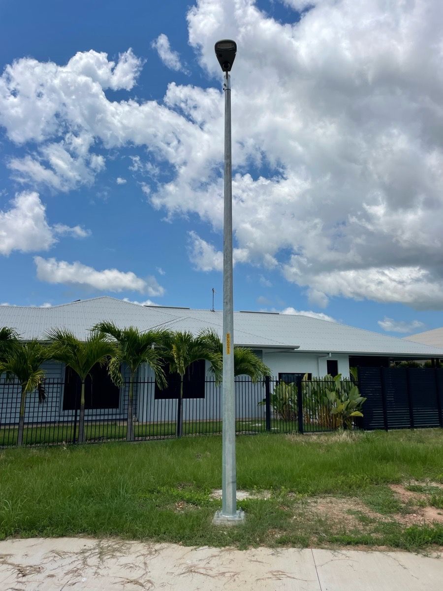Tall white light pole in a grassy yard beside a fence and low building under a cloudy blue sky