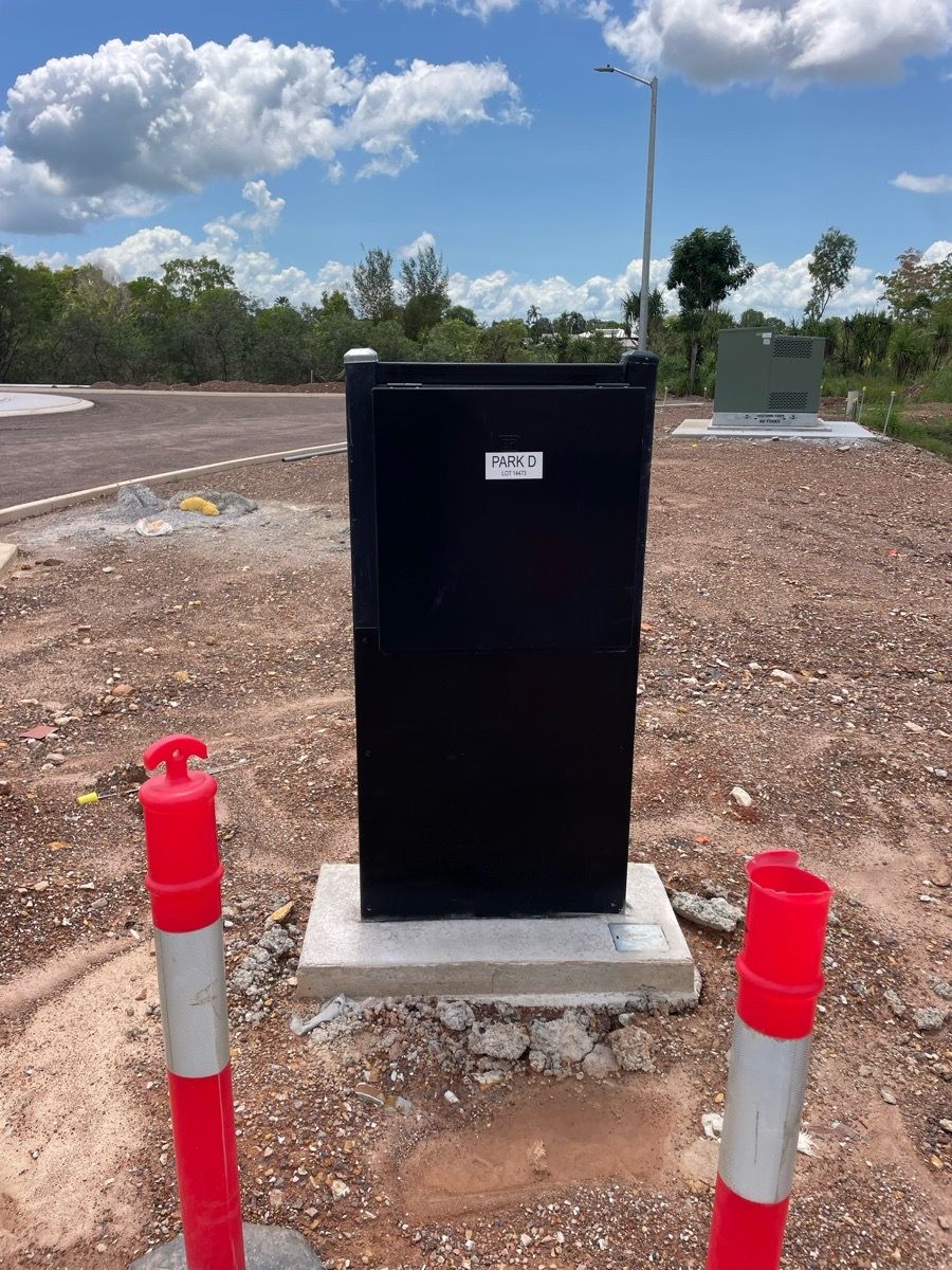 Black utility box on concrete pad beside two red-and-white bollards in a gravel lot under a blue sky.
