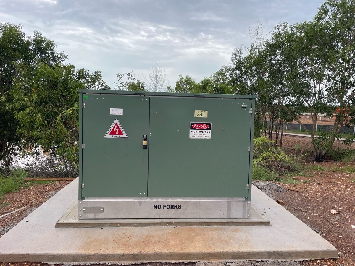Green electrical utility box on a concrete pad, surrounded by trees and brush outdoors.