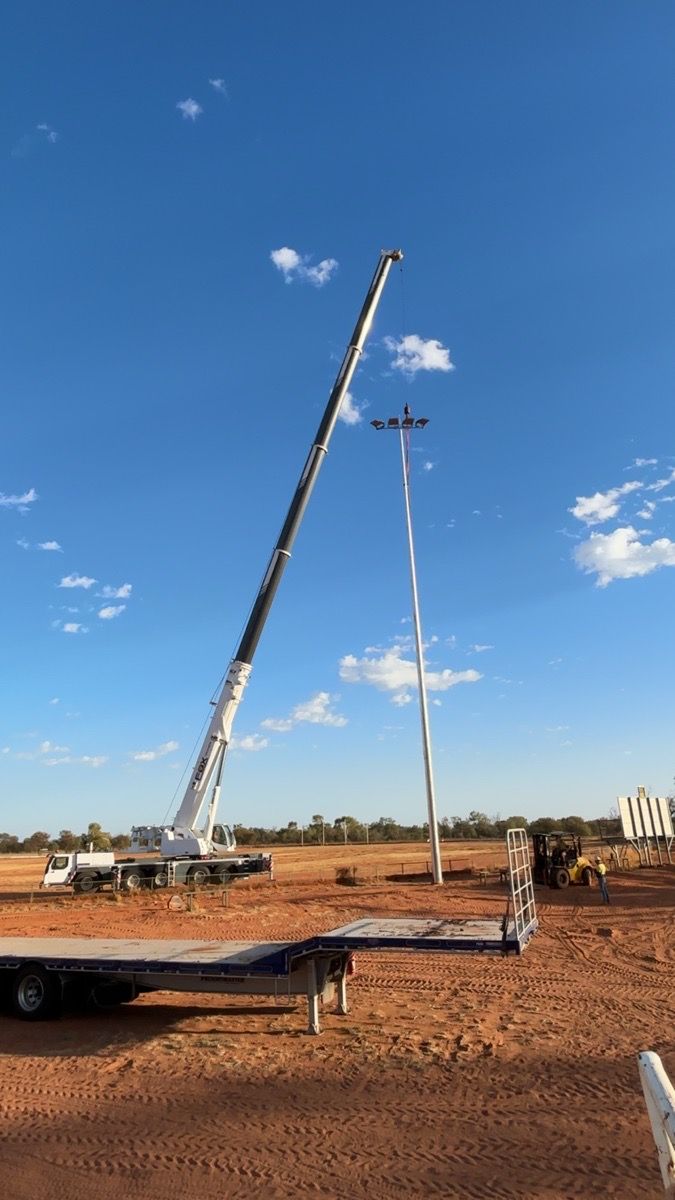 Construction crane lifting a tall utility pole at a dusty site under a clear blue sky.
