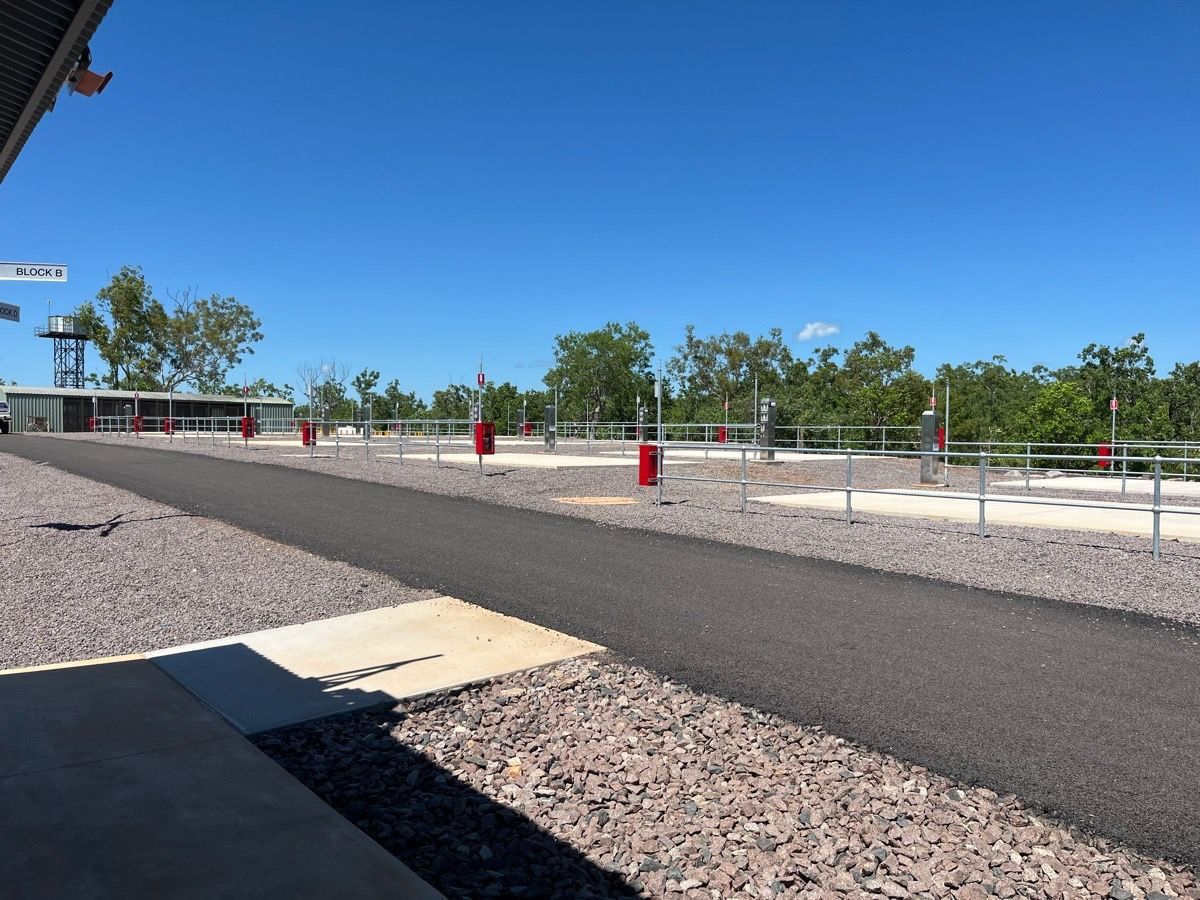 Empty paved path beside a gravel lot under a clear blue sky, with red posts and trees in the distance