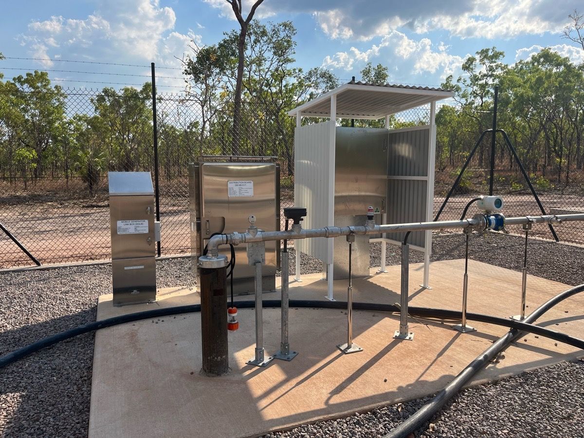Outdoor utility equipment and pipes on a gravel pad beside a fenced field, under a small metal shelter.