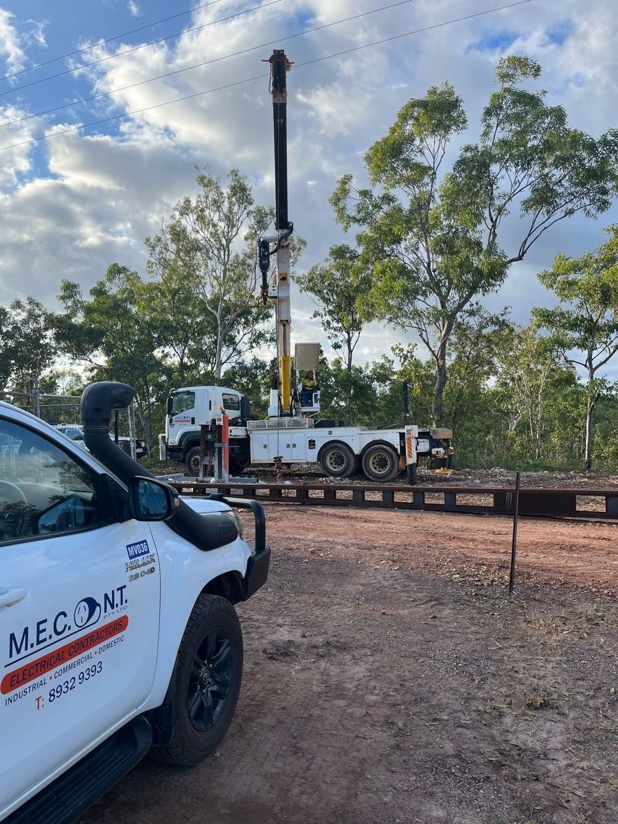 White utility truck with crane beside a flatbed trailer in a dirt lot, trees and cloudy sky in back.