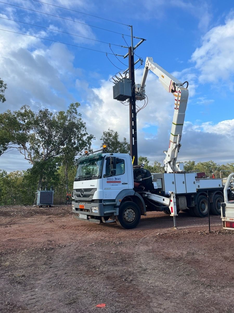 White utility truck with raised boom working beside a utility pole on a dirt roadside under a blue sky.