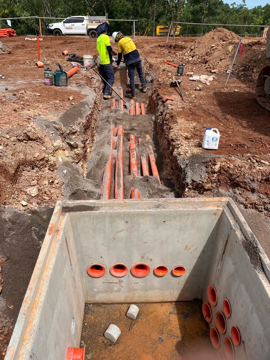 Construction workers installing orange utility pipes in a trench beside a concrete manhole.