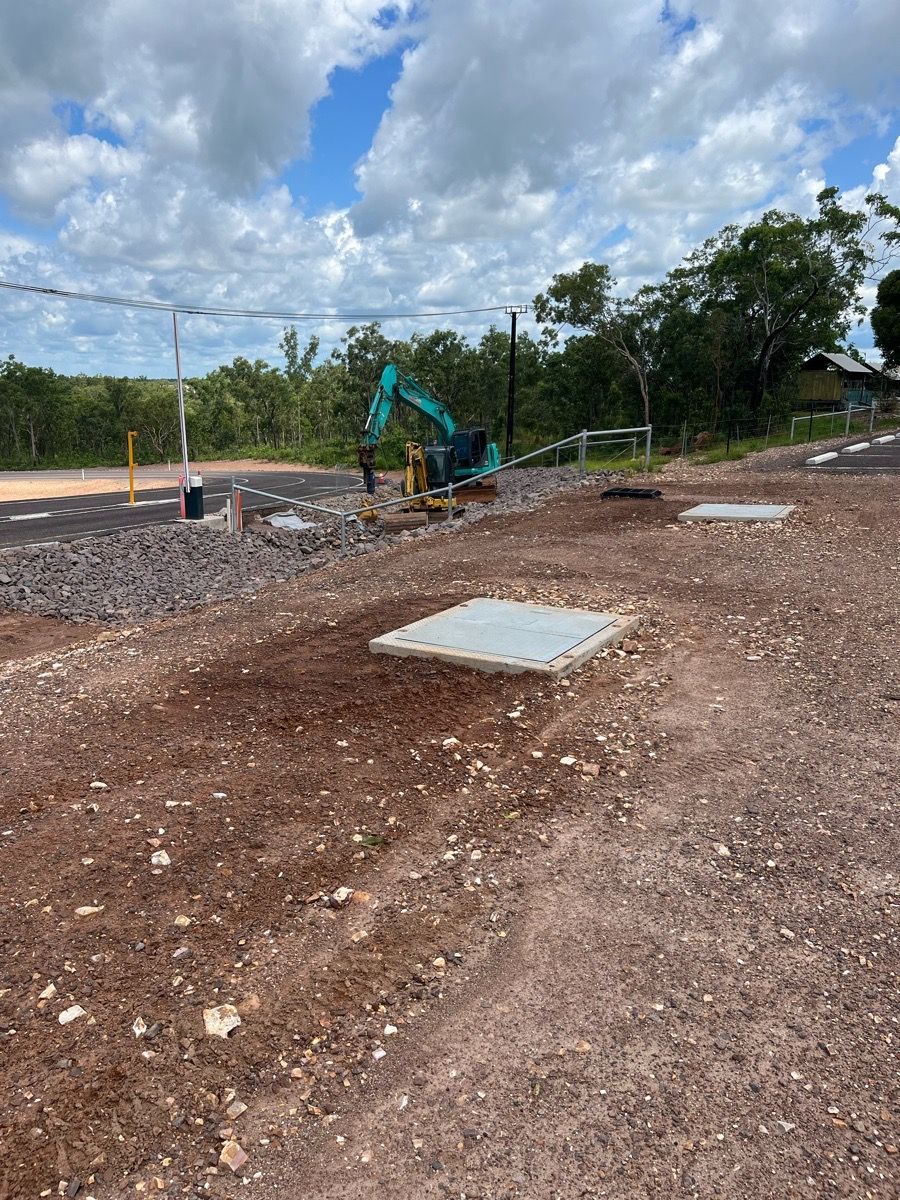 Construction site with excavator on dirt road, concrete slabs, and trees under a cloudy sky