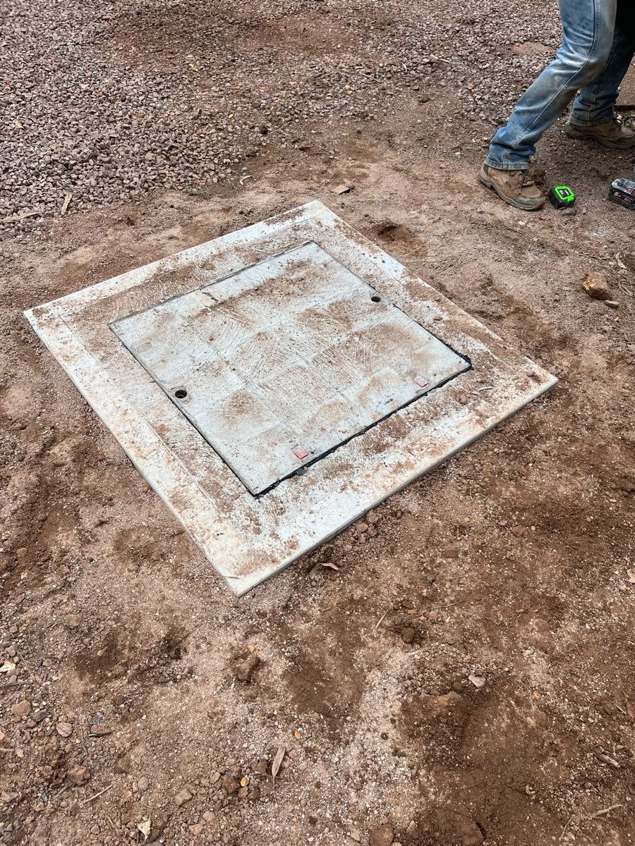 Concrete utility access cover set in muddy ground, with a person's boots visible at the edge.