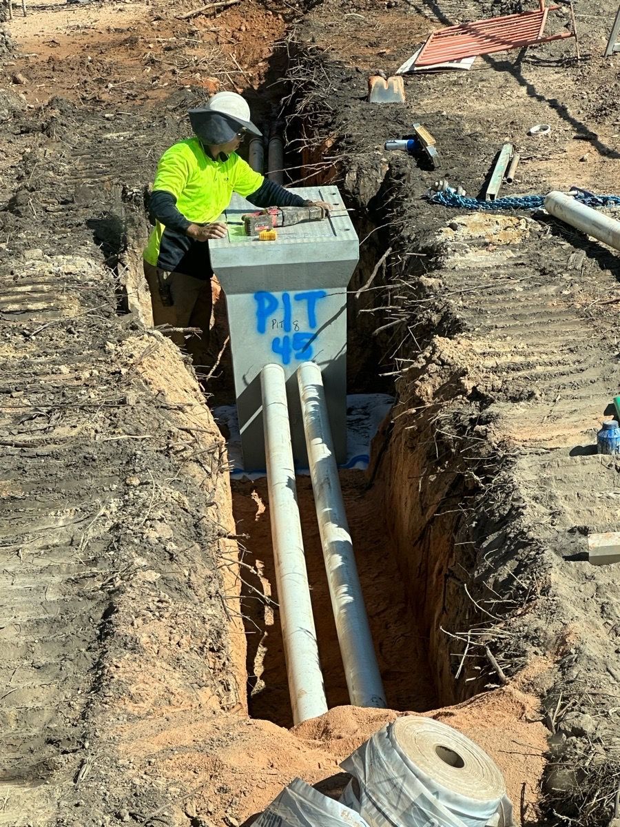 Worker in neon safety shirt standing in a trench beside exposed utility pipes and a concrete utility box.