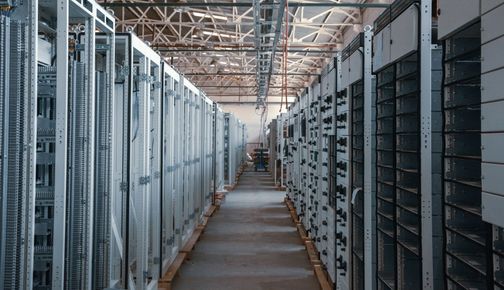 Rows of industrial server racks in a data center aisle with overhead piping and ceiling lights