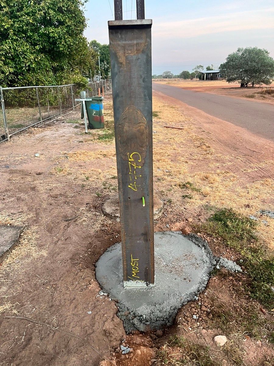 Concrete utility pole beside a dirt road, set in a roadside base with trees in the background.
