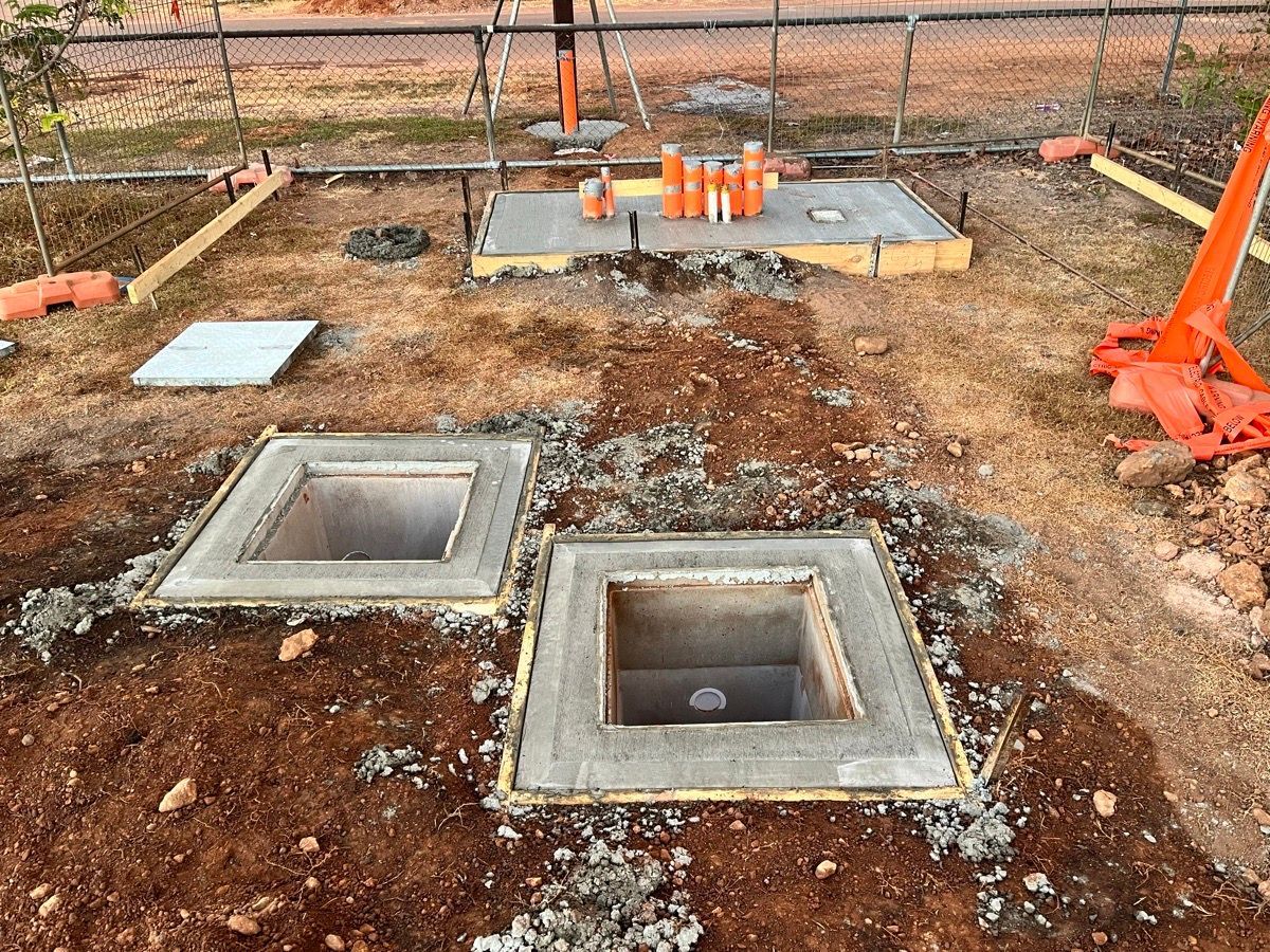 Construction site with two rectangular utility pits, orange equipment, and dirt ground near a fence
