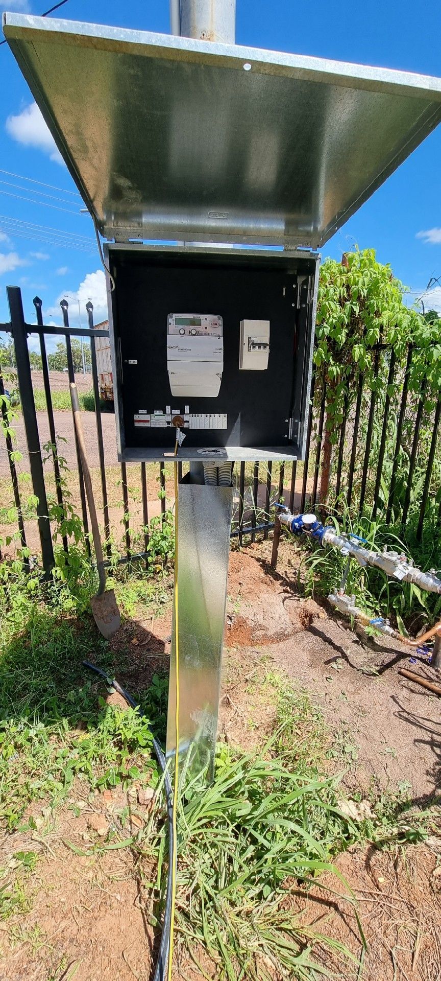 Outdoor utility box on a metal post with an open lid beside a fence and plants