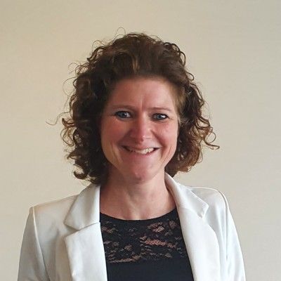 Vrouw met krullend bruin haar, gekleed in een witte blazer, lacht naar de camera.