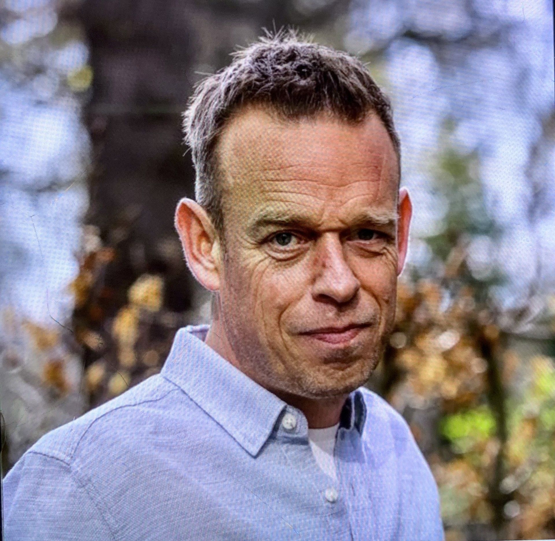 Een man in een lichtblauw overhemd kijkt buiten in de camera. Op de achtergrond zijn bomen te zien.