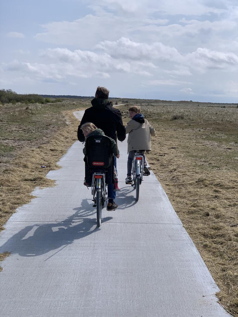 Een persoon fietst met twee kinderen over een verhard pad door een grasrijk landschap, met een bewolkte hemel erboven.