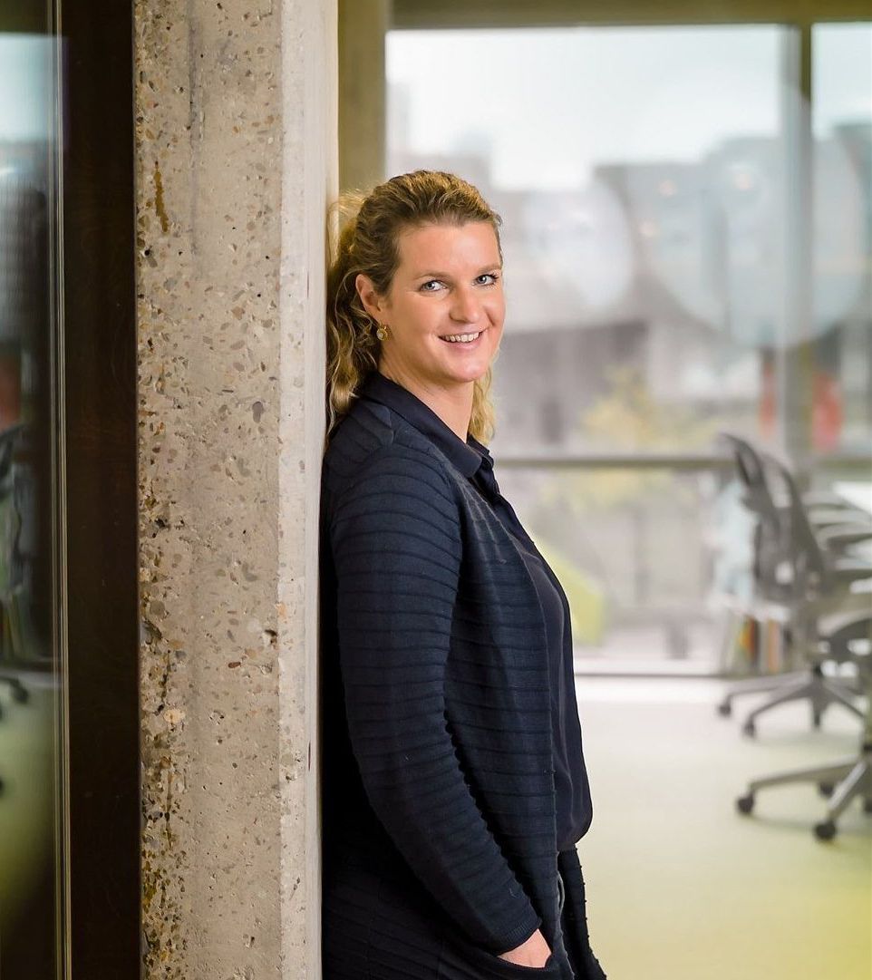 Vrouw leunt tegen een betonnen pilaar en lacht. Ze draagt een donkerblauw vest en bevindt zich in een kantooromgeving met een wazige achtergrond.