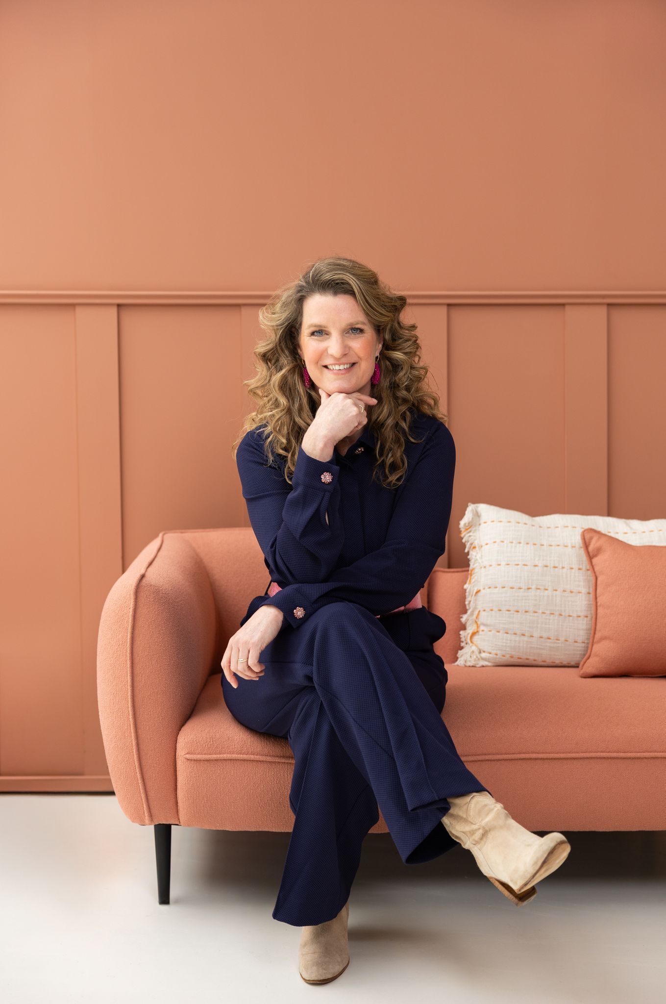 Een vrouw in een blauw pak zit glimlachend op een perzikkleurige bank voor een perzikkleurige muur.