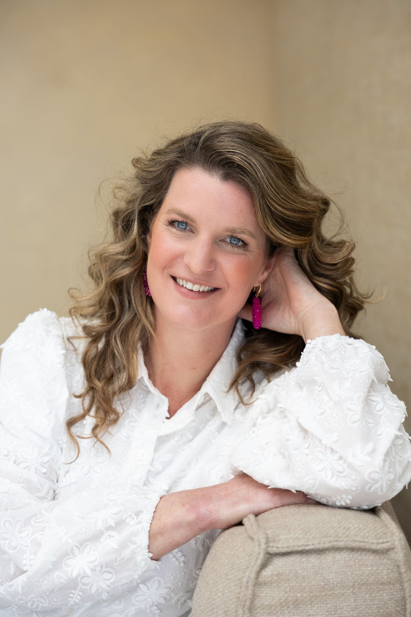 Vrouw in witte blouse glimlacht, haar arm rustend op de bank. Roze oorbellen.