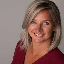 Vrouw met blond bobkapsel, lachend, draagt een rode blouse, tegen een grijze achtergrond.