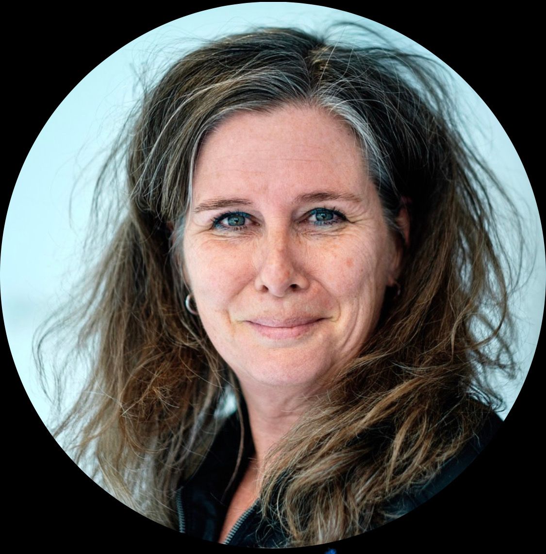 Vrouw met lang, golvend bruin haar en blauwe ogen, lachend, voor een lichte achtergrond.
