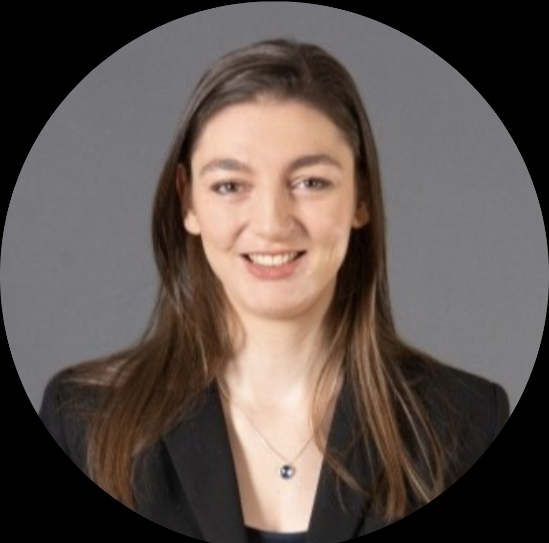 Vrouw met lang bruin haar lacht, draagt ​​een donkere blazer en een ketting en staat voor een grijze achtergrond.