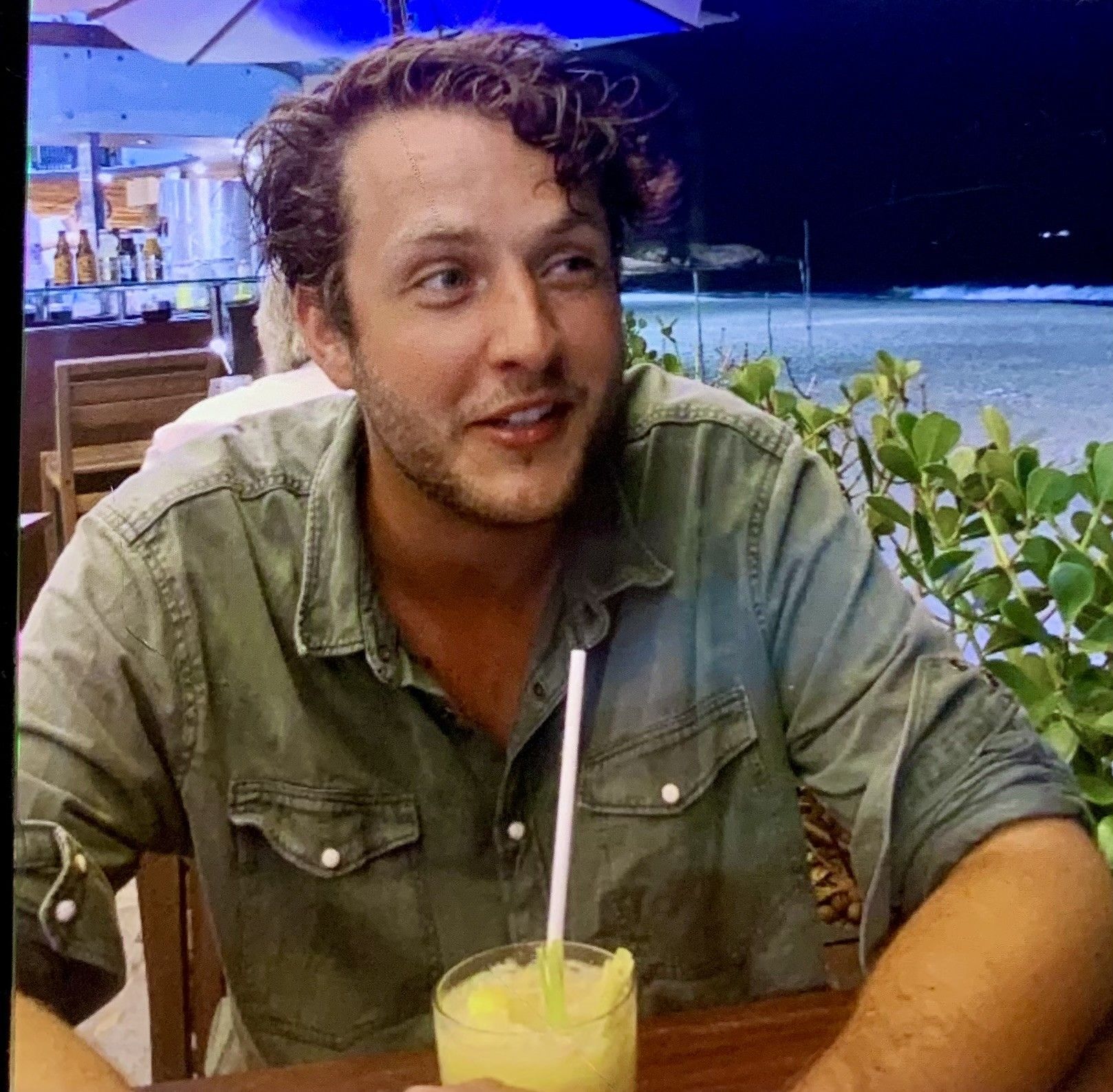 Een man met krullend haar in een groen shirt zit aan een tafeltje buiten, vlakbij de oceaan, en geniet van een drankje.