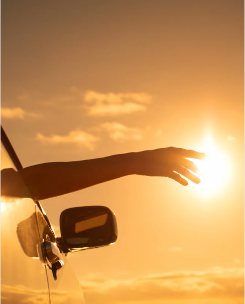 Een arm die uit het autoraam steekt, gericht naar de felle zon, een warme, gouden hemel.