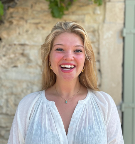 Een blonde vrouw, gekleed in een witte blouse, staat lachend voor een stenen muur.